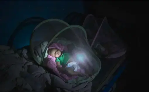 An infant wearing a green shirt and diapers sleeps in a dark room on a pink blanket covering some kind of infant bed. The bed has a canopy of mesh fabric.
