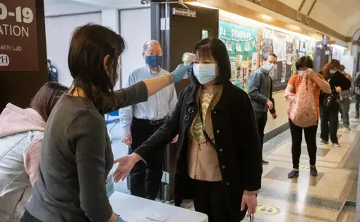 A woman wearing a mask stands in front of a table as the first person in a long line of people. She is holding out her hand to one woman behind the table as another woman behind the table takes her temperature using an infrared forehead thermometer.