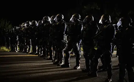 Police officers in riot gear line up across a street.