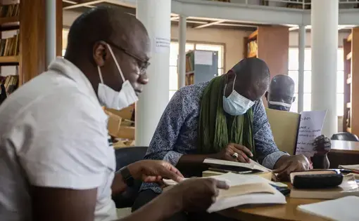 Three people wearing medical face masks sit and read papers and books in an archive.