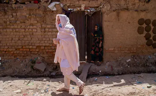 A woman is walking toward the left of the frame. She's wearing clear plastic gloves, a hospital mask pulled down around her chin, a light pink scarf covering her head and draped around her body. Behind her, another woman stands in the doorway of a stone building.