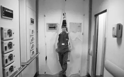 Black and white photo of a man wearing scrubs and a white face mask walking through white curtains that have a sign on them saying "keep out."