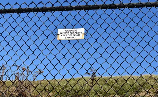 Image of a black chain-link fence with a sign that reads "Warning, private property, keep off fence, bad dog!"