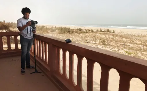 A person stands on a balcony with a camera on a monopod.
