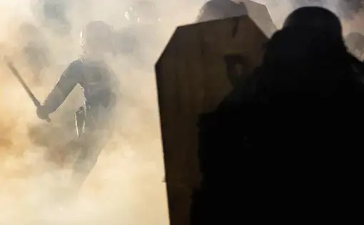 In the background, a police officer wields a club against a protester amidst a cloud of teargas. In the foreground, someone's back is to the camera, and they are holding a shield.