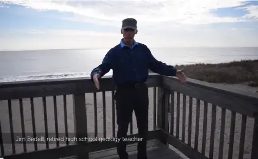 A man wearing a blue button-down shirt and brown cap stands on a deck in front of the beach.