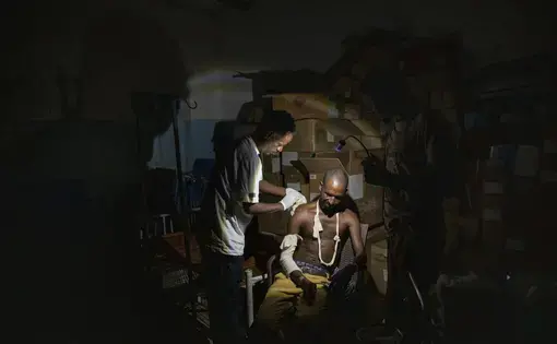 A doctor treats a wound on a seated man's shoulder. A third man points a small lamp at the two in order to illuminate them in the otherwise dark room. Behind them is a tower of cardboard boxes.