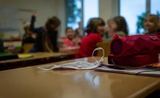 In the foreground, a white face mask sits on a school desk along with a pencil case and other supplies. In the blurred background, a group of young schoolchildren sit and talk together in a classroom.