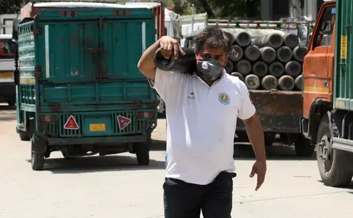 A man carries an oxygen cylinder. There are trucks in the background.