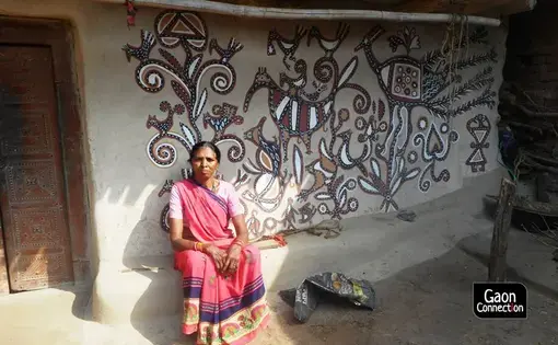 An older Indian woman in a pink sari sits in front of a wall painted with a somewhat abstract black, brown, and white mural featuring animals.