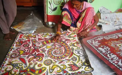 An older Indian woman in a pink sari squats next to a large square paper painted with a semi-abstract red, yellow, and black pattern. The corner of a similar painting is visible to the right of the frame.