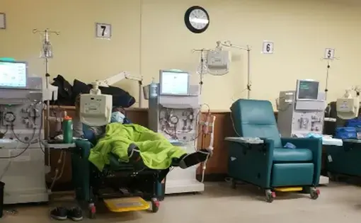 Image of a patient covered in a lime green blanket reclining in a dark green chair. The patient is wearing a blue hospital mask and is hooked up to a machine. Also visible are two other machines and another chair, which is vacant.