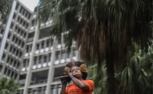 A man in traditional Indigenous dress stands in front of a tall building and plays a flute-like instrument.