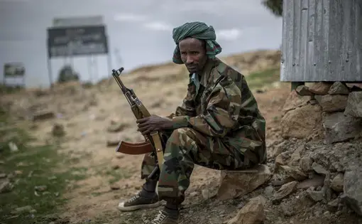 Image of a Tigray soldier wearing camo sits on a rock on the ground. He's holding a rifle and wears a green scarf on his head.