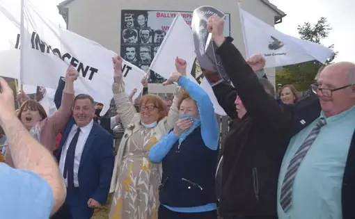 People stand and cheer in front of a building. Some hold white flags that read 'Innocent.'