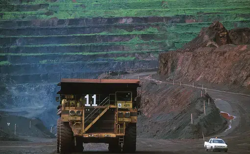 a truck drives on a dirt road with an open mine behind it