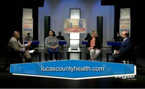Image of six people sitting on a stage with their chairs spaced several feet apart. At the bottom of the frame, a chyron reads "lucascountyhealth.com."