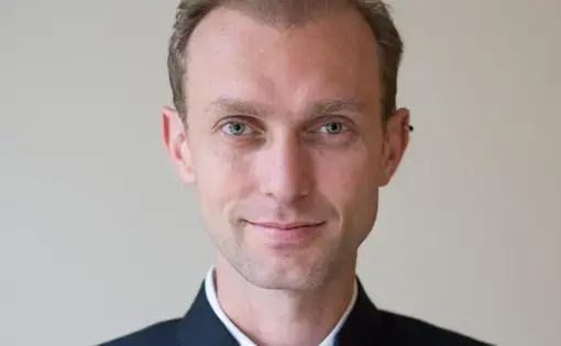 A man wearing a white shirt and dark blue jacket. He has an ear piercing.