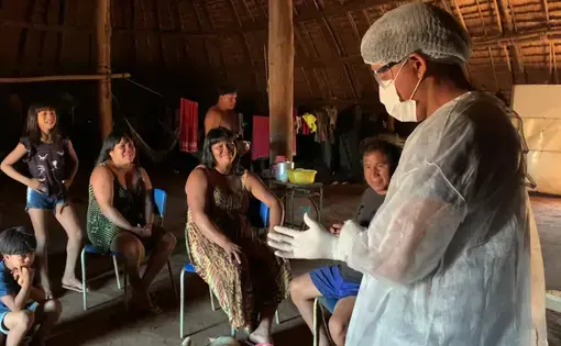 A person wearing personal protective equipment (PPE) gear is talking to Indigenous Xingu individuals. In the foreground, there are people sitting on chairs.