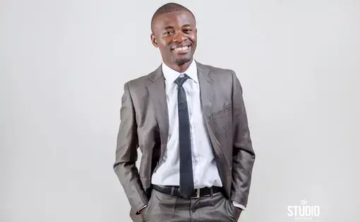 A person wearing a grey jacket, white shirt, and black tie smiles at the camera.