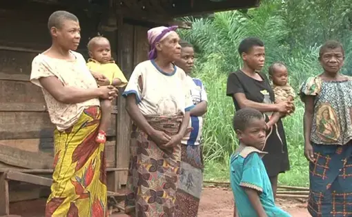 Six people stand in the foreground of a wooden house. They are looking at the camera.
