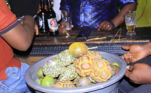 A person holds a plastic blue basket of fruits. Two other people sit nearby.