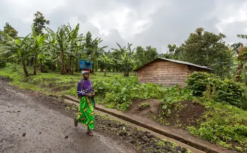 A woman carries a basket on her head as she walks by a wooden house.