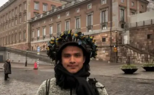 A person wears a grey winter jacket and an Indigenous headdress. In the foreground is a building.