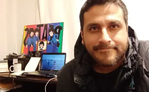 A person wears a black jacket. In the foreground, there is a study desk with a laptop and a poster board.