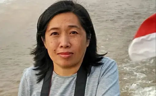 A person wears a grey long sleeve shirt and is carrying a camera. In the foreground there is a beach with ocean waves.