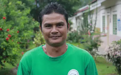 Image of a person wearing a green shirt. A house and tropical forest is in the background.