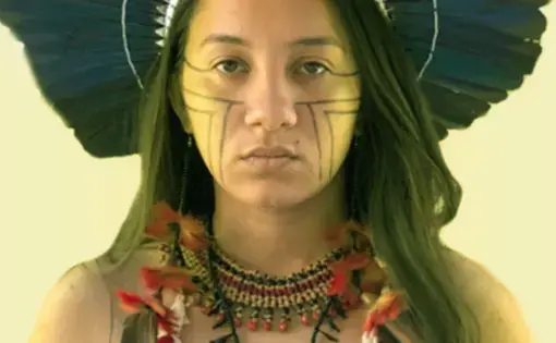 A person with black hair and Indigenous headdress looks into the camera.