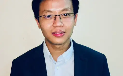 A person with a black jacket, light blue shirt, and glasses.
