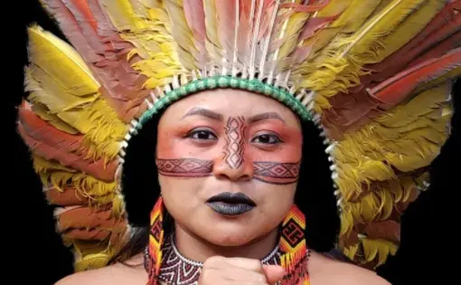 A woman with red makeup, yellow shirt and indigenous headress stares at the camera