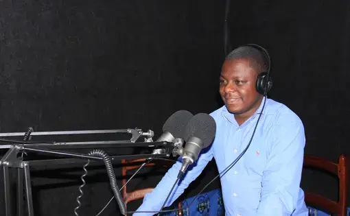A person wears a blue shirt and stands over a microphone. He is also wearing headphones and is in a radio station.