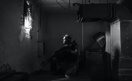 A person holds a gun and sits in the middle of a dark room, looking toward a small window.