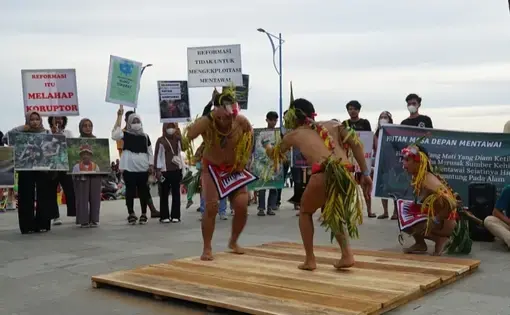 Two people dress in Indigenous clothing and dance on a wooden platform in front of an audience.
