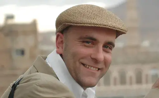 A man wears a brown hat, white scarf, and a brown jacket.
