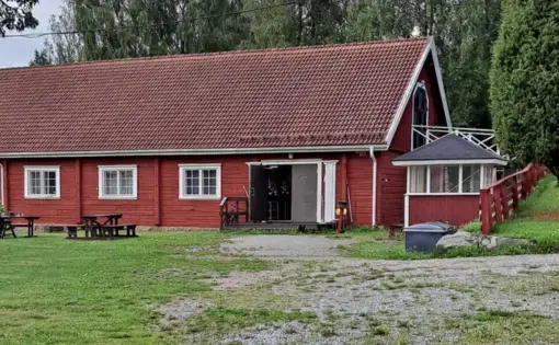 a red wooden house on a grassy lot surrounded by trees, picnic tables