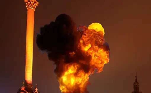 an explosion billows above a building beside a plaza, lighting up a cloudy nigh sky in orange. a statue in the foreground.