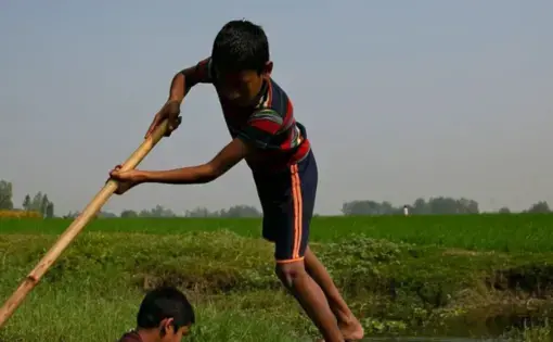 one boy pushes a boat while standing in the water while another uses a rod to redirect the small wooden vessel.