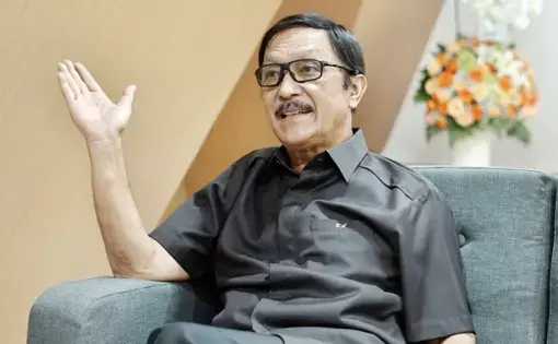 A man wears a black shirt and black eyeglasses. He raises his hand while talking.