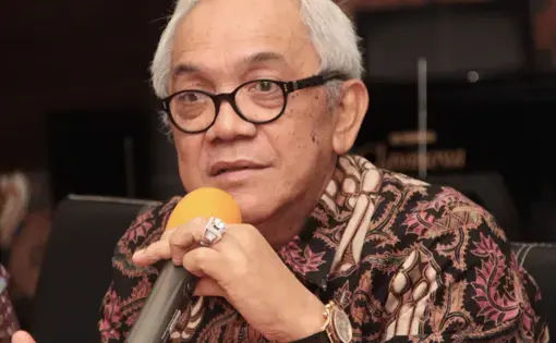 A man wears a brown, batik-patterned shirt and black eyeglasses while talking with a mic.