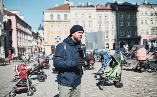 Journalist with microphone stands in a plaza filled with empty strollers
