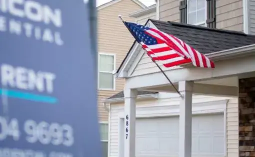 a rental sign from Tricon Residential hangs in the yard of a two story house where an American flag flies on the front porch.