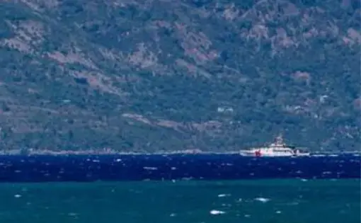 the photo is taken from the water, looking towards land. In the distance, close to the shore, a white boat patrols the water