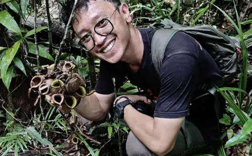 A man wears a black shirt and trekking pants. He has eyeglasses and is holding a plant.
