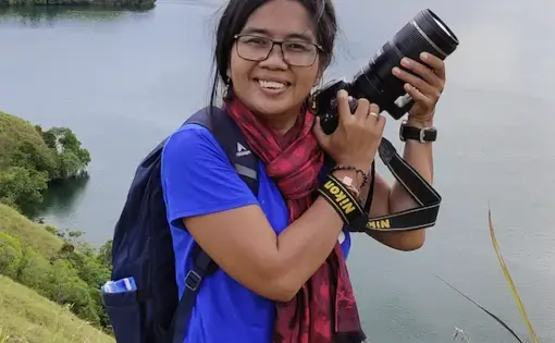 A woman holds a camera in her hands. She wears glasses, a purple shirt and jeans.