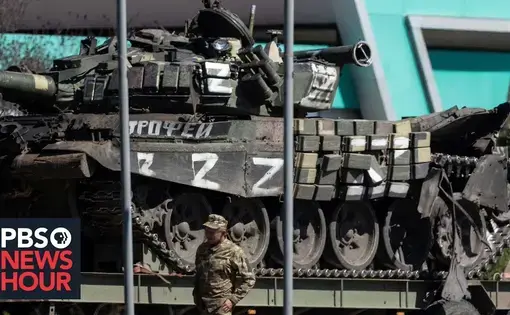 a soldier stands next to a trailer loaded with a military tank with Cryllic markings
