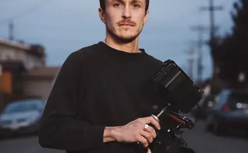 Headshot of Brandon Tauszik. He wears a black long-sleeved crewneck sweatshirt and holds a video camera.